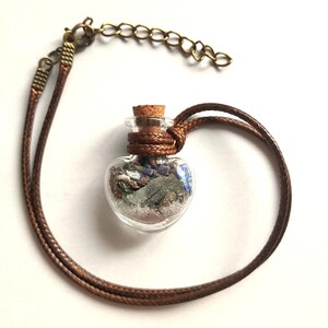 May include: A heart-shaped glass vial pendant filled with dried herbs, small stones, and sand, sealed with a cork stopper. The vial is suspended from a brown cord necklace featuring a bronze-toned metal chain extender and lobster clasp.