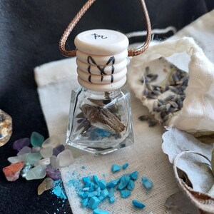 May include: A small, clear, faceted glass bottle with a light-colored, ribbed wooden cap featuring a hand-drawn black symbol and the letters 'tn'. The bottle contains dried botanical elements and is suspended by a thin, brown cord.