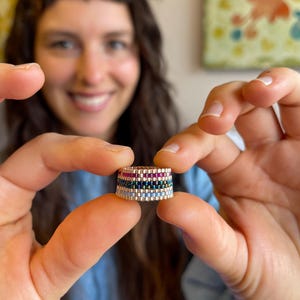Puede incluir: Un anillo elaborado con pequeñas cuentas rectangulares dispuestas en filas horizontales. El diseño presenta una fila superior de cuentas rosa vibrante, una fila central de cuentas verde oscuro y una fila inferior de cuentas azul claro.
