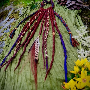 May include: A set of deep red and blue synthetic dreadlocks featuring decorative accents including light-colored thread wraps, wooden beads, and a single brown and white striped feather. The dreadlocks are arranged in a fan-like pattern.