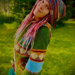 May include: A multi-colored striped knit beanie with a small rectangular tag and a long-sleeved green knit sweater featuring a geometric diamond pattern in brown, cream, and blue, finished with a cream-colored fringe along the bottom hem.