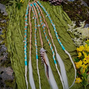 May include: A set of light brown and white synthetic hair extensions attached to a circular loop. The strands are braided and wrapped with bright turquoise cord, featuring small metallic charms, beads, and a single patterned feather accent.