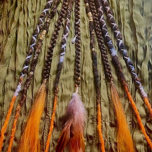 May include: A set of brown hair extensions featuring multiple braids adorned with metallic beads, light blue yarn wraps, and a central feather accent. The extensions transition into loose, vibrant orange hair strands at the bottom ends.