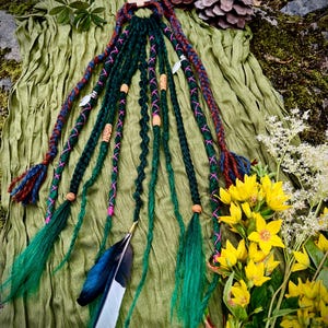 May include: A set of dark green synthetic dreadlocks featuring pink thread wraps, wooden beads, and silver-toned charms. The hair extensions are accented with a large black and white feather and include braided sections with multi-colored fiber ends.