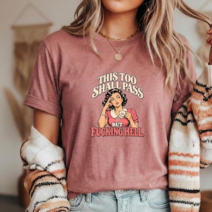May include: A mauve short-sleeved t-shirt featuring a graphic of a person holding a teacup with the text "THIS TOO SHALL PASS BUT FUCKING HELL." The design includes a retro-style illustration of a person with dark hair wearing a pink top.