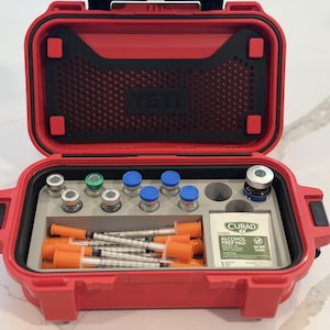 May include: A red YETI hard-shell case with an open lid, containing a grey foam insert holding several small glass vials with colored caps, multiple syringes with orange caps, and a single Curad alcohol prep pad in a white square packet.