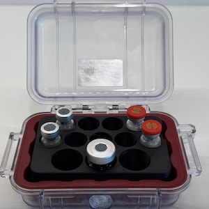 May include: A clear, rectangular, hard-shell protective case with a hinged lid and a red interior seal. Inside, a black foam insert with twelve circular cutouts holds five small glass vials with silver and red caps in various positions.