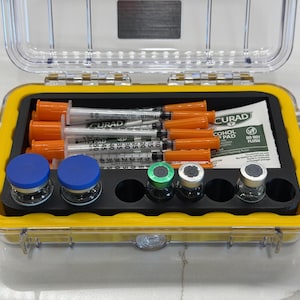 May include: A clear, rectangular protective case with a yellow interior seal containing medical supplies. Inside are several syringes with orange caps, small glass vials with blue, green, and silver caps, and a Curad alcohol prep pad.