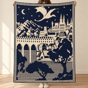 May include: A rectangular crochet blanket featuring a dark navy blue and cream color scheme. The design depicts a fantasy landscape with a flying dragon, a crescent moon, stars, a mountain range, a castle, and an arched stone bridge.