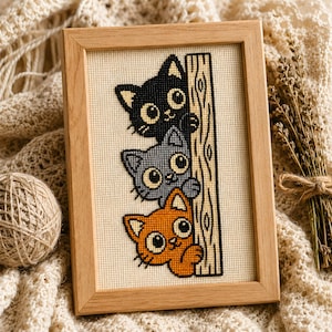 May include: A framed cross-stitch embroidery featuring three stacked cats in black, grey, and orange, each with large eyes, peeking from behind a vertical wooden post. The design is stitched on cream-coloured Aida fabric within a light wood frame.