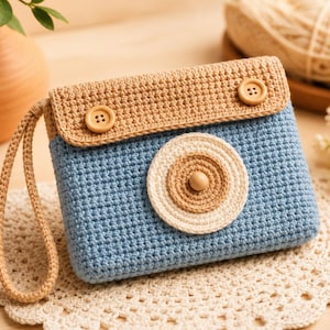 May include: A rectangular crochet pouch in light blue and tan yarn, designed to resemble a camera. It features a tan flap with two wooden buttons, a circular lens detail in tan and cream, and a matching tan crochet wrist strap attached to the side.