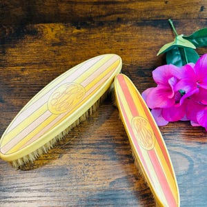 May include: Two oval-shaped brushes with light-colored bristles, featuring a yellow and peach striped pattern on the back. Each brush is decorated with a circular gold-toned monogram emblem centered on the top surface of the handle.