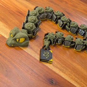May include: An articulated snake toy in olive green with bright yellow eyes. The snake features a series of connected segments, each displaying a small shield emblem. A black rectangular padlock with a yellow snake graphic hangs from the tail end.