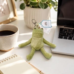 May include: A plush, light green frog keychain with a soft, textured fabric surface. The frog has a simple, rounded head, two small black eyes, and outstretched limbs. A silver metal lobster clasp is attached to the top, along with a small white tag.