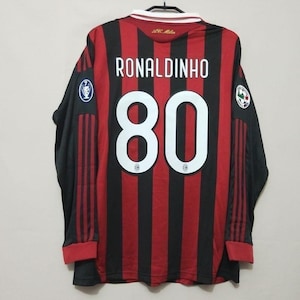 Puede incluir: Una camiseta de fútbol del AC Milan de manga larga con rayas verticales rojas y negras. La parte posterior muestra el nombre RONALDINHO y el número 80 en letras blancas. Incluye un cuello blanco, parches en las mangas y el logo del A.C. Milan.