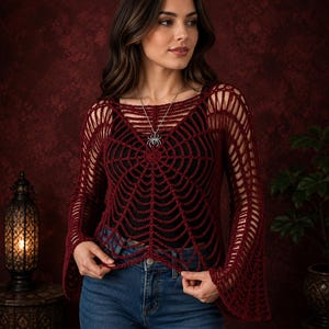 Puede incluir: Una blusa de crochet de manga larga en color rojo oscuro con un patrón de telaraña en la parte delantera, combinada con un collar de araña de plata, una camiseta negra debajo y pantalones vaqueros azules. La prenda tiene mangas acampanadas.