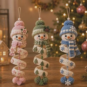 Puede incluir: Tres adornos colgantes de muñeco de nieve hechos a ganchillo con cuerpos en espiral. El muñeco de la izquierda lleva un gorro y bufanda rosa con flores. El del centro lleva un gorro y bufanda verde con árboles. El de la derecha lleva un gorro y bufanda azul con copos de nieve.