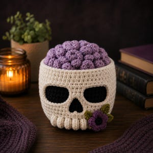 Puede incluir: Un recipiente con forma de calavera tejido a ganchillo en hilo color hueso con cuencas oculares y cavidad nasal negras. La parte superior está rellena de formas de hilo morado redondeadas y texturizadas que parecen un cerebro con una flor.