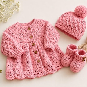 Puede incluir: Un cárdigan de ganchillo rosa con cinco botones marrones, un gorro a juego con un pompón y un par de patucos de ganchillo rosas. Un pequeño conejo de ganchillo beige está colocado junto a los patucos, completando este conjunto de artículos hechos a mano.
