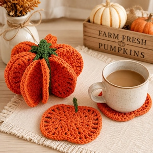 Puede incluir: Un juego de posavasos de ganchillo con forma de calabaza naranja y tallos verdes. Un posavasos está apilado en forma de calabaza redondeada, mientras que otro yace plano debajo de una taza de cerámica moteada con una bebida marrón claro.