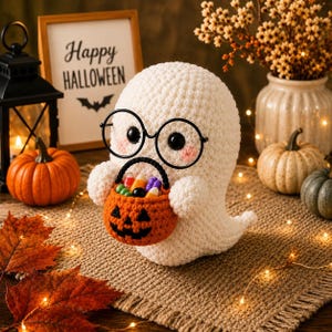 Puede incluir: Una figura de fantasma tejida a ganchillo en color blanco, con gafas redondas negras y sosteniendo un pequeño cubo de calabaza naranja tejido a ganchillo lleno de dulces. El fantasma tiene mejillas rosadas y ojos de cuentas negras.