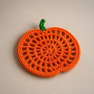 May include: A round, orange crochet coaster shaped like a pumpkin, featuring an intricate openwork pattern throughout the body and a small, textured green stem attached to the top edge, creating a flat, circular textile design.