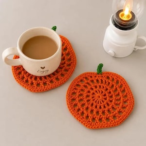 May include: Two round orange crochet coasters shaped like pumpkins with small green stems. One coaster supports a beige ceramic mug filled with coffee, featuring the word Enjoy and a small heart icon printed on the front surface.