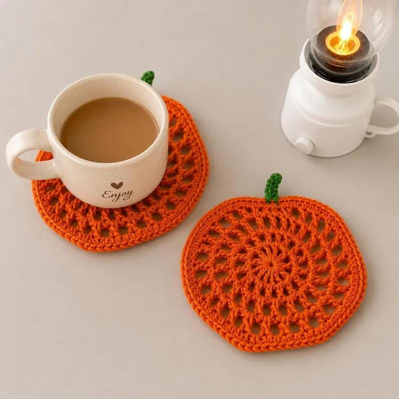 May include: Two round orange crochet coasters shaped like pumpkins with small green stems. One coaster supports a beige ceramic mug filled with coffee, featuring the word Enjoy and a small heart icon printed on the front surface.