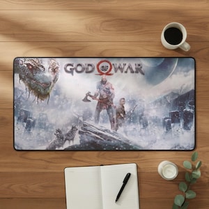 Może przedstawiać: Prostokątna podkładka na biurko z grafiką z gry wideo God of War. Projekt przedstawia centralną męską postać trzymającą topór, młodszego towarzysza, głowę wielkiego węża oraz wilki w zaśnieżonym krajobrazie z napisem God of War.