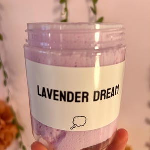 May include: A clear plastic jar filled with a light purple, textured substance. The jar features a white label with the black text "LAVENDER DREAM" and a small black outline of a thought bubble icon printed below the text.