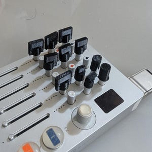 May include: A silver audio mixing controller featuring a series of black 3D-printed knobs attached to metallic shafts. The device includes vertical sliders, a large circular knob with a white center, and two round buttons with orange and blue inserts.
