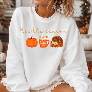 Könnte beinhalten: Ein weißes Langarm-Sweatshirt mit dem Text 'tis the season' in orangefarbener Schreibschrift. Unter dem Text befinden sich drei Illustrationen: ein leuchtend orangefarbener Kürbis, eine orangefarbene Tasse mit der Aufschrift 'Pumpkin spice' und ein Igel im Herbstlaub.
