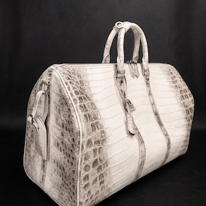 Peut inclure: Un sac de voyage blanc et gris avec un motif texturé en peau de crocodile. Le sac comprend deux poignées supérieures, une fermeture éclair sur le dessus avec deux tirettes et une étiquette de bagage suspendue sur le côté avec une boucle.