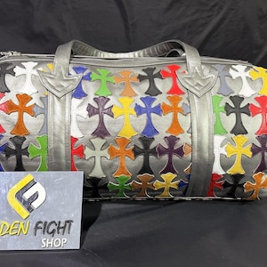 May include: A silver duffel bag featuring a repeating pattern of multi-colored leather cross appliques in red, blue, yellow, green, orange, purple, black, and white. The bag has two top handles and a zipper closure. A sign reads GOLDEN FIGHT SHOP.