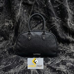 May include: A black leather handbag featuring a rounded top, two sturdy handles, and a series of embossed cross motifs across the front. A small silver-toned metal plaque is centered at the bottom. The bag is displayed against a dark textured fabric.