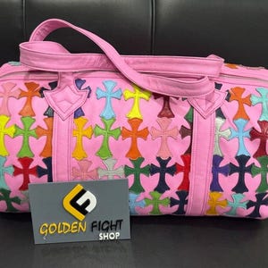 May include: A pink leather duffel bag featuring a repeating pattern of multi-colored cross-shaped patches in yellow, green, orange, blue, and red. The bag has two top handles and a grey rectangular sign with the text GOLDEN FIGHT SHOP.