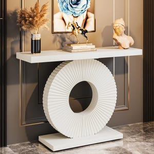 May include: A white console table featuring a rectangular top and base connected by a large, circular, vertically ribbed pedestal. The table surface holds a black vase with dried grass, a stack of books, a horse figurine, and a bust sculpture.