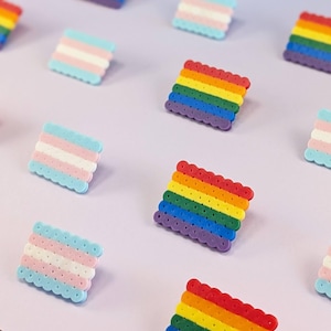 May include: A collection of square pins made from small fused plastic beads. Some pins feature horizontal stripes in red, orange, yellow, green, blue, and purple, while others feature horizontal stripes in light blue, pink, and white colors.