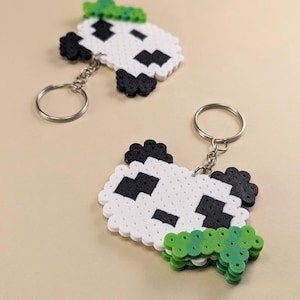 May include: Two panda-shaped keychains crafted from white, black, and green plastic fuse beads. Each keychain features a circular metal ring attached by a short chain to the pixelated panda head, which is accented with a green leaf-like element.