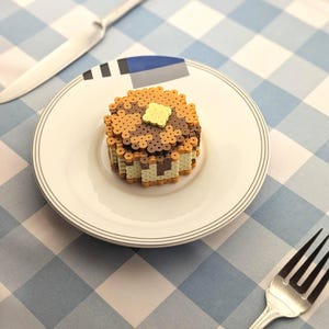 May include: A three-dimensional pancake stack crafted from small, circular plastic beads in shades of brown, tan, and cream, topped with a single square yellow butter pat, resting on a white ceramic plate with a thin gray rim and blue accents.