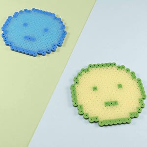 May include: Two circular coasters crafted from fused plastic beads. One coaster is light blue with a darker blue border and a simple face design. The second coaster is pale yellow with a green border and a matching simple face design.