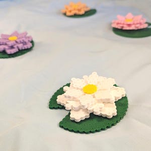 May include: Four water lily flowers crafted from small plastic beads, each resting on a green circular lily pad. The flowers are white, pink, purple, and peach, each featuring a bright yellow center. The beads create a textured, pixelated appearance.