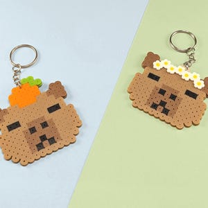 May include: Two capybara keychains made from brown, tan, and black fuse beads. One keychain features a bright orange carrot on the head, while the other features a white and yellow daisy flower crown. Both include a silver metal split ring.