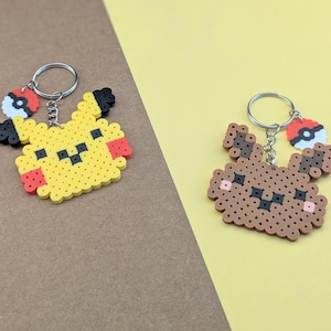 May include: Two pixel art keychains made from fused plastic beads. One keychain features a yellow character with black ear tips and red cheeks, while the other is a brown character with dark brown ear accents and pink cheeks. Both include a small Poké Ball charm.