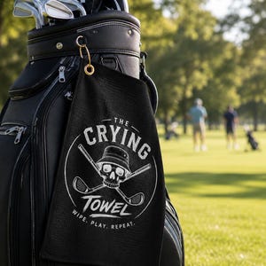May include: A black waffle-textured golf towel hanging from a black golf bag. The towel features a white circular graphic with a skull wearing a bucket hat, crossed golf clubs, and the text: THE CRYING TOWEL WIPE. PLAY. REPEAT. The towel is attached with a gold ring.