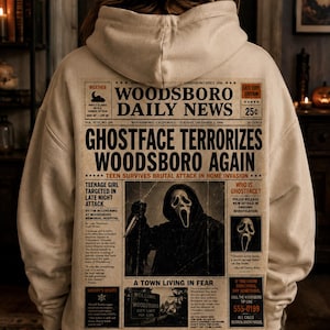May include: A beige hooded sweatshirt featuring a large graphic on the back designed like a newspaper front page titled Woodsboro Daily News. The print includes the headline Ghostface Terrorizes Woodsboro Again and an image of the masked figure.
