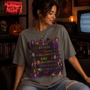 May include: A grey short-sleeve t-shirt featuring a graphic print of stacked VHS tapes labeled with horror movie titles including Beetlejuice, A Nightmare on Elm Street, Halloween, Saw, Hocus Pocus, Edward Scissorhands, The Exorcist, and IT.