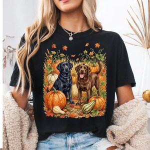 Peut inclure: Un t-shirt noir à manches courtes avec une impression graphique représentant un Labrador noir et un Labrador chocolat assis parmi des citrouilles orange, des courges et des feuilles d'automne. Les chiens portent des colliers.