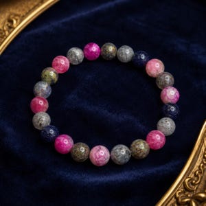 Può includere: Un braccialetto circolare composto da perline sferiche lisce in una varietà di colori, tra cui rosa vibrante, blu profondo con macchioline scintillanti, grigio screziato e toni verdi terrosi. Le perline sono disposte su un cordino flessibile.