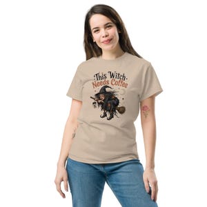 May include: A beige short-sleeve t-shirt featuring a graphic of a witch flying on a broomstick while holding a coffee cup. The text above the illustration reads This Witch Needs Coffee in black and orange lettering, accompanied by small star and moon icons.